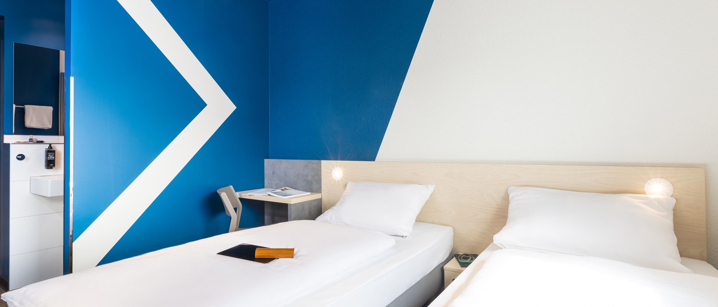 Modernes Zimmer mit zwei Einzelbetten, blauer Wand und kleinem Schreibtisch. Minimalistisches Design mit geometrischen Mustern., © DQuadrat Living GmbH Modernes Zimmer mit zwei Einzelbetten, blauer Wand und kleinem Schreibtisch. Minimalistisches Design mit geometrischen Mustern., © DQuadrat Living GmbH