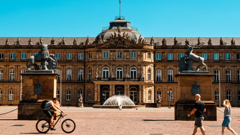 The New Palace in Stuttgart in sunny weather. People walking and a cyclist riding in the foreground. Two statues flank the entrance., © SMG Stuttgart Marketing GmbH - Sarah Schmid