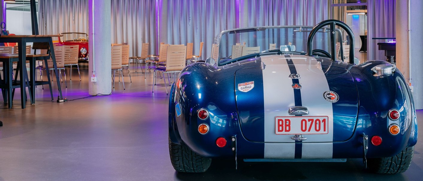 Ein blauer Oldtimer mit weißen Streifen steht in einem modernen Tagungsraum mit Tischen und Stühlen., © V8 HOTEL