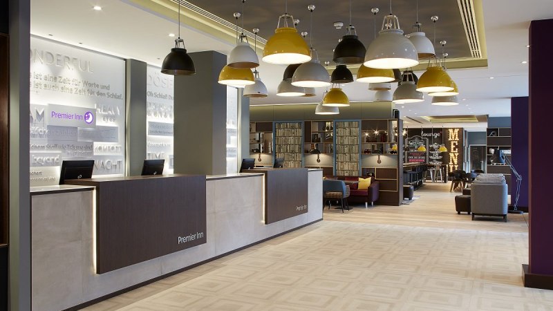 A modern hotel lobby with a front desk, hanging lamps, and a cozy seating area. "Premier Inn" is written on the wall. A lounge is visible in the background., © Premier Inn A modern hotel lobby with a front desk, hanging lamps, and a cozy seating area. "Premier Inn" is written on the wall. A lounge is visible in the background., © Premier Inn