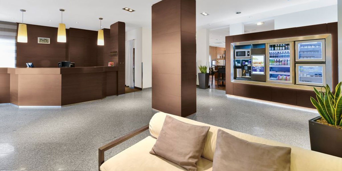 Moderne Lobby mit brauner Rezeption, beigem Sofa, Snackautomaten und Pflanzen. Helle Beleuchtung und glänzender Boden., © NH Stuttgart Sindelfingen Moderne Lobby mit brauner Rezeption, beigem Sofa, Snackautomaten und Pflanzen. Helle Beleuchtung und glänzender Boden., © NH Stuttgart Sindelfingen
