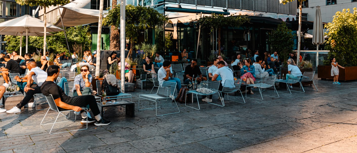 Menschen genießen das sonnige Wetter in einem belebten Außenbereich eines Cafés. Moderne Gebäude und Bäume umgeben die Szene., © SMG, Sarah Schmid