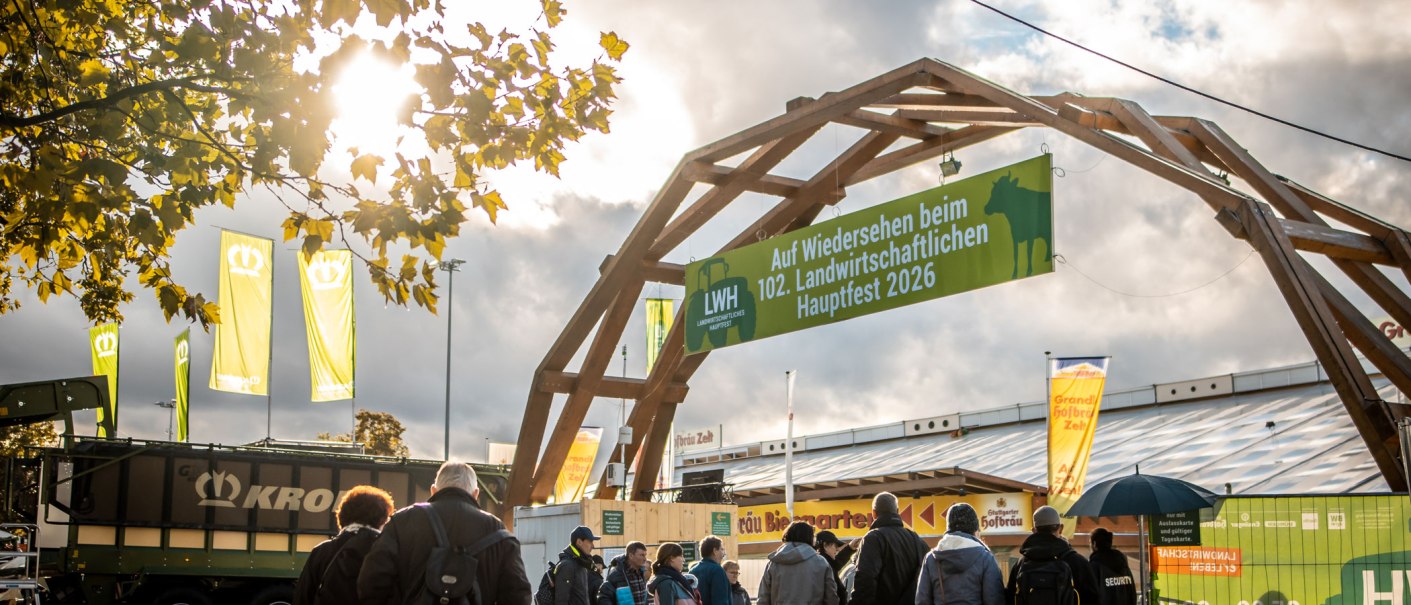 Besucher betreten das Landwirtschaftliche Hauptfest 2026 unter einem Holzbogen mit Banner. Herbstliche Atmosph&auml;re mit gelben Bl&auml;ttern und Wolken., &copy; Daniel Schneider, 2022