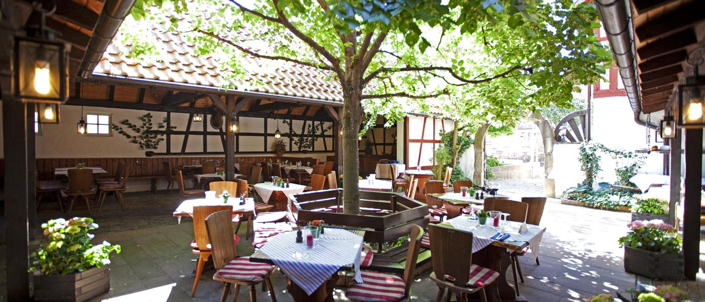 Ein gemütlicher Innenhof mit Tischen und Stühlen unter einem Baum, umgeben von Fachwerkhäusern. Die Tische sind mit gestreiften Tischdecken gedeckt., © Hotel Hirsch