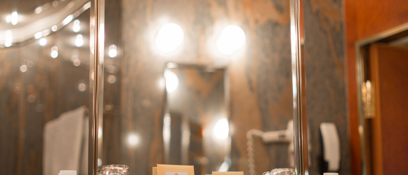 Luxuriöses Badezimmer mit großen Spiegeln, heller Beleuchtung und Pflegeprodukten auf der Ablage. Marmorierte Wände und moderne Armaturen., © Hotel Royal