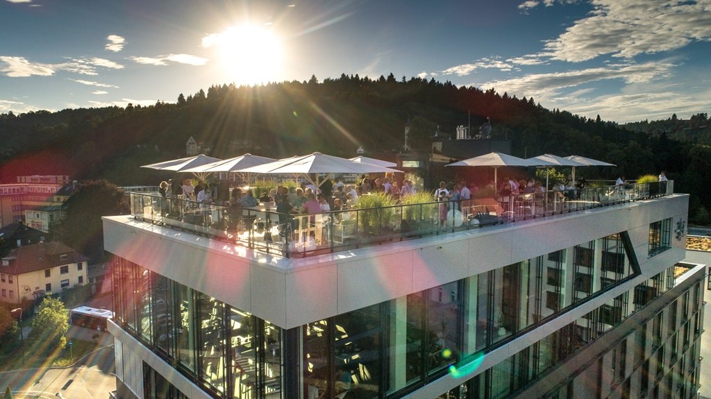 Modern roof terrace with people under parasols at sunset, surrounded by forest and buildings., © Hotel am Remspark Modern roof terrace with people under parasols at sunset, surrounded by forest and buildings., © Hotel am Remspark