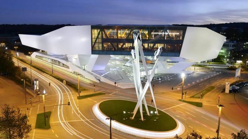 The Porsche Museum at night, illuminated with modern architecture and a sculpture of three cars on high pillars., © Porsche Museum The Porsche Museum at night, illuminated with modern architecture and a sculpture of three cars on high pillars., © Porsche Museum
