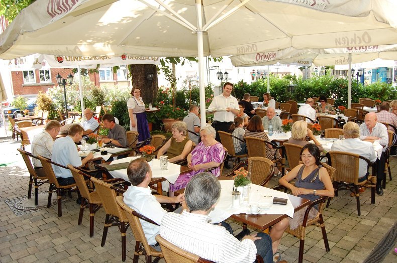 A bustling beer garden filled with people sitting at tables under large umbrellas. Waiters are serving the guests. It’s a sunny day., © Jeanette Bender A bustling beer garden filled with people sitting at tables under large umbrellas. Waiters are serving the guests. It’s a sunny day., © Jeanette Bender
