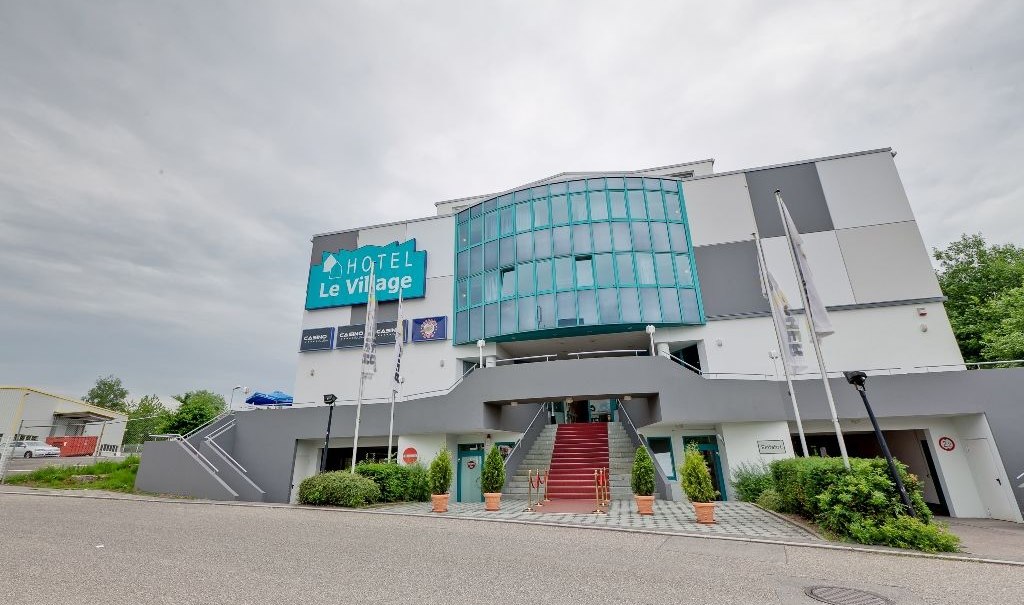 Das Hotel Le Village zeigt eine moderne Fassade mit Glasfront und Treppe. Vor dem Gebäude wehen Fahnen, der Himmel ist bewölkt., © Hotel Le Village