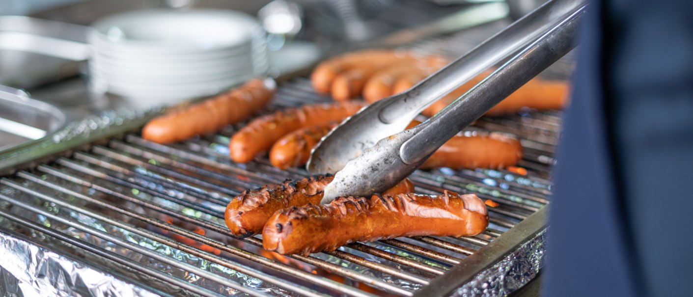 W&uuml;rstchen werden auf einem Grill mit einer Zange gewendet. Im Hintergrund sind unscharfe Teller zu sehen., &copy; Frank Nonnenmann