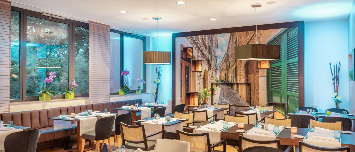 Elegantes Restaurant mit modernen Tischen, St&uuml;hlen und Wandbild einer Gasse. Dezente Beleuchtung und dekorative Pflanzen schaffen eine gem&uuml;tliche Atmosph&auml;re., &copy; NH Hotel Stuttgart Airport