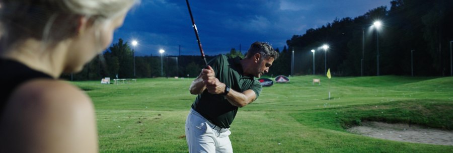 Ein Mann beim Golfschwung auf einem beleuchteten Platz in der Dämmerung, beobachtet von einer Frau im Vordergrund., © GolfKultur Stuttgart