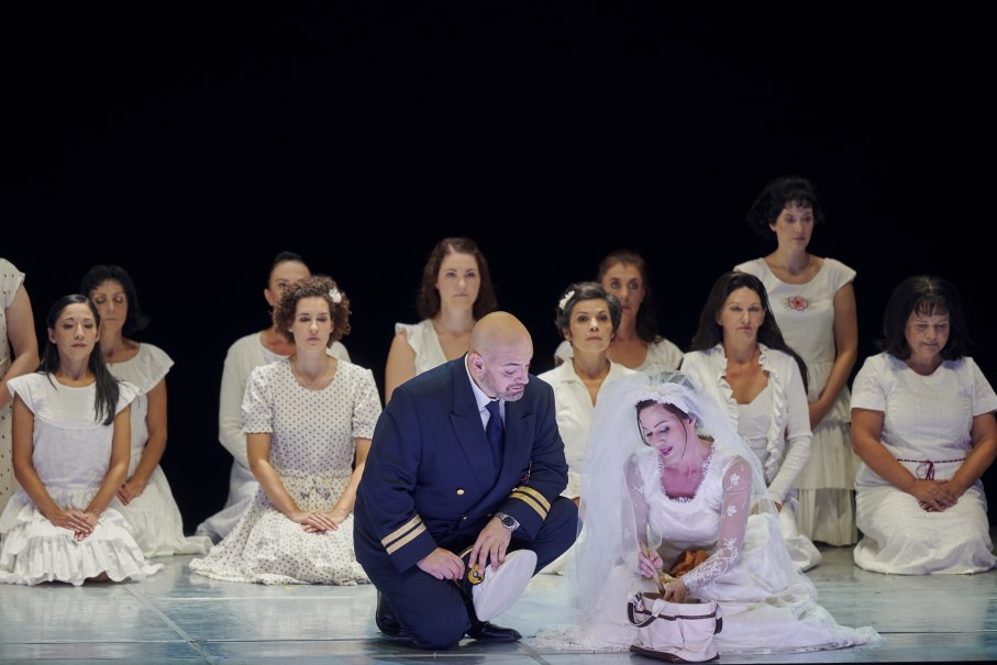 Ein Mann in Uniform kniet neben einer Frau im Brautkleid. Um sie herum sitzen Frauen in weißen Kleidern. Die Szene wirkt dramatisch und emotional., © Martin Sigmund
