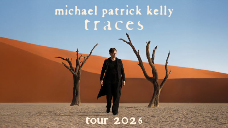 Ein Mann in schwarzer Kleidung steht in einer W&uuml;stenlandschaft mit kahlen B&auml;umen und rotem Sand. Der Himmel ist blau. Text: Michael Patrick Kelly - Traces Tour 2026., &copy; links im Bild