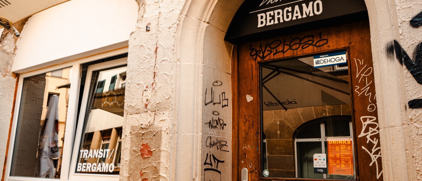 Eingang mit der Aufschrift 'Transit Bergamo' über der Tür, umgeben von Graffiti. Ein Fenster zeigt Spiegelungen der Umgebung., © SMG_Sarah Schmid
