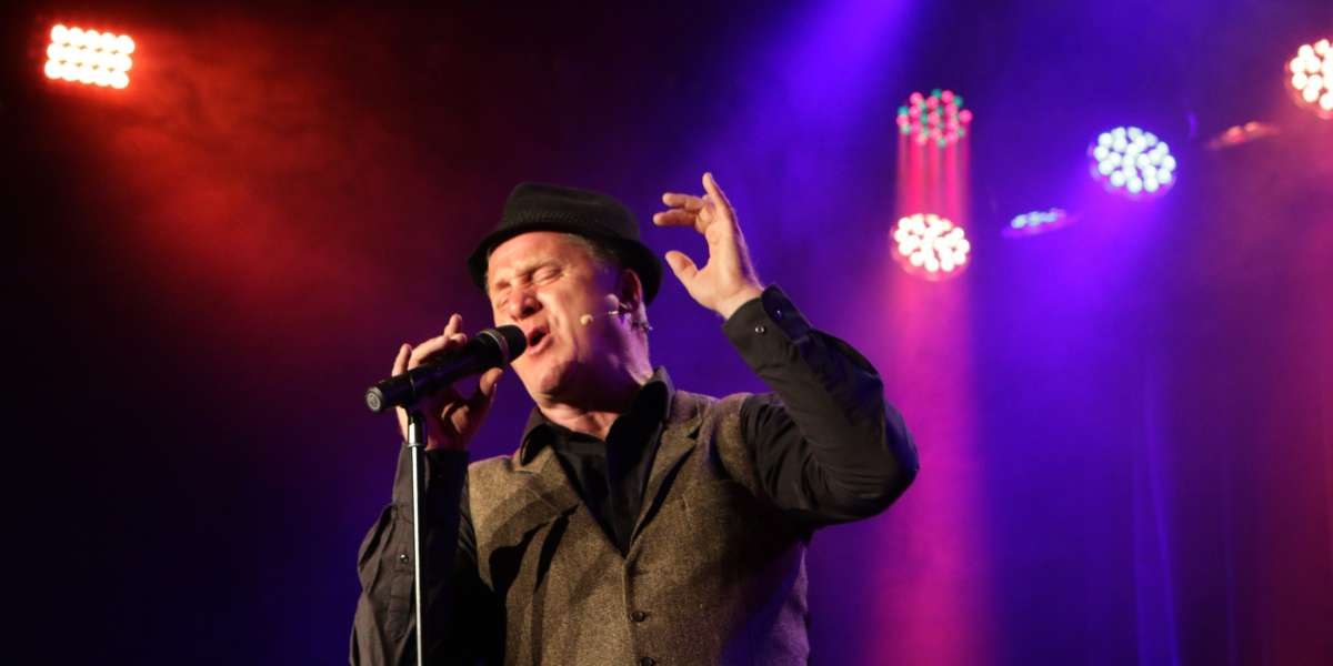 A singer in a hat performs passionately on a stage surrounded by colorful lights., © Sabine Haymann