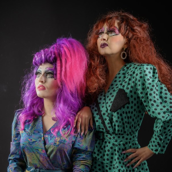 Zwei Drag Queens in bunten, auffälligen Outfits und extravagantem Make-up posieren selbstbewusst vor einem dunklen Hintergrund., © Veranstalter Zwei Drag Queens in bunten, auffälligen Outfits und extravagantem Make-up posieren selbstbewusst vor einem dunklen Hintergrund., © Veranstalter