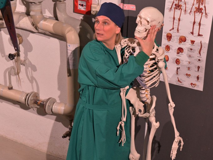 Person in gr&uuml;ner Kleidung umarmt ein Skelett in einem Raum mit medizinischen Postern und Rohren an der Wand., &copy; Landesmuseum W&uuml;rttemberg