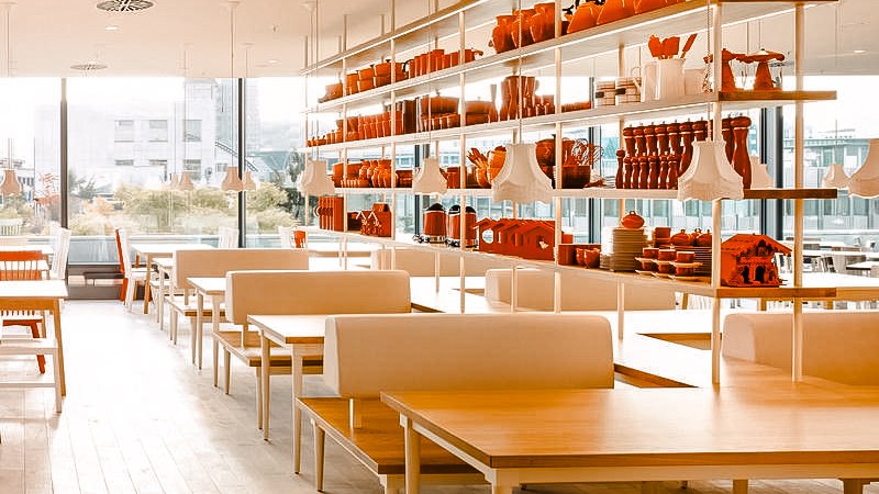 Modernes Restaurant mit weißen Tischen und Regalen voller roter Dekorationen. Große Fenster bieten Blick nach draußen., © Breuninger Modernes Restaurant mit weißen Tischen und Regalen voller roter Dekorationen. Große Fenster bieten Blick nach draußen., © Breuninger