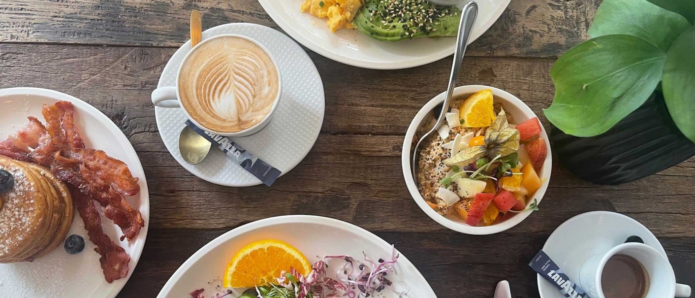 Ein reich gedeckter Frühstückstisch mit Kaffee, Pancakes mit Speck, Avocado mit Rührei und einer Schale mit frischem Obst., © SMG Ein reich gedeckter Frühstückstisch mit Kaffee, Pancakes mit Speck, Avocado mit Rührei und einer Schale mit frischem Obst., © SMG