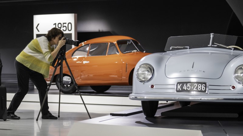 Eine Person fotografiert ein blaues Porsche-Auto in einem Museum. Im Hintergrund steht ein orangefarbenes Auto. Ein Koffer steht neben der Person., © Porsche AG Eine Person fotografiert ein blaues Porsche-Auto in einem Museum. Im Hintergrund steht ein orangefarbenes Auto. Ein Koffer steht neben der Person., © Porsche AG