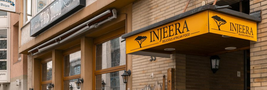 Restaurant Injeera with yellow sign and wooden fence, offers African cuisine. Exterior view with lanterns and plants., © SMG Stuttgart Marketing GmbH - Sarah Schmid