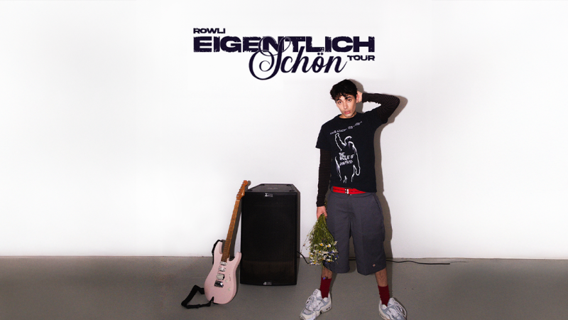 Ein junger Mann mit Blumen in der Hand steht neben einer Gitarre und einem Verstärker. Im Hintergrund ist der Schriftzug 'Rowli - Eigentlich schön Tour 2026' zu sehen., © links im Bild Ein junger Mann mit Blumen in der Hand steht neben einer Gitarre und einem Verstärker. Im Hintergrund ist der Schriftzug 'Rowli - Eigentlich schön Tour 2026' zu sehen., © links im Bild