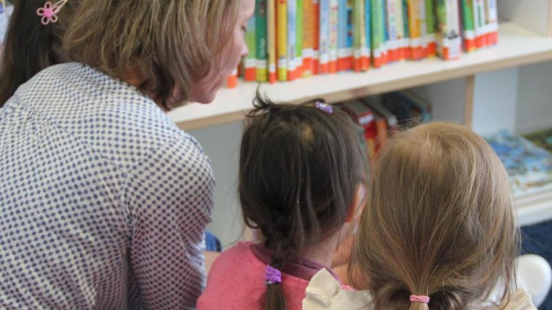 Eine Frau sitzt mit drei Kindern vor einem Bücherregal und liest ihnen vor. Die Kinder hören aufmerksam zu., © Stadtbibliothek Stuttgart - Leseohren e.V.