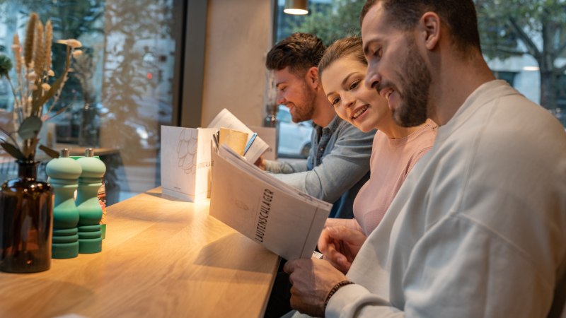 Three people are sitting at a table in a restaurant reading the menu. In the background is a window with a view outside., © Stuttgart-Marketing GmbH, Martina Denker