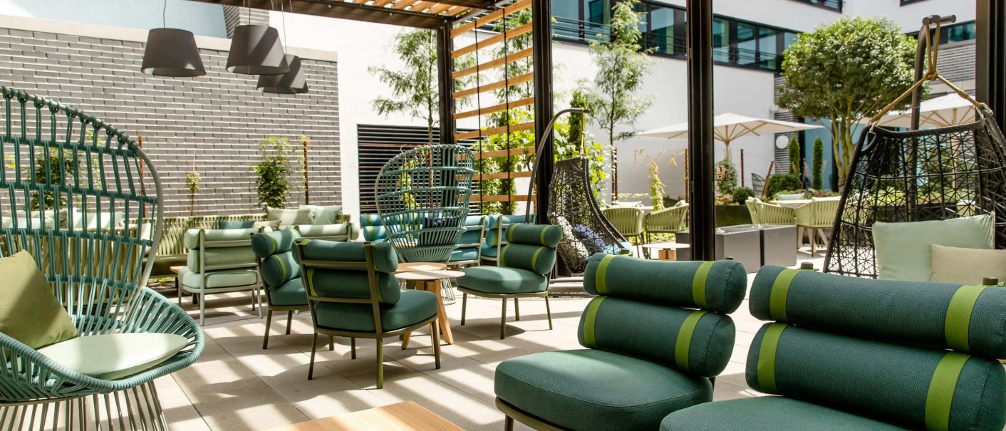 Moderne Terrasse mit grünen Sitzmöbeln, Holzpergola und Pflanzen. Hängesessel und Tische schaffen eine einladende Atmosphäre im Freien., © Motel One - Stuttgart Hauptbahnhof
