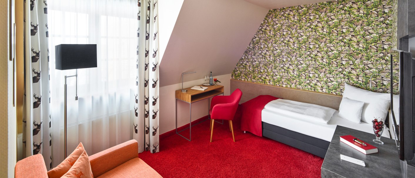 Ein Einzelzimmer mit rotem Teppich, Einzelbett, Schreibtisch und Sessel. Die Wand hat ein Steinmuster, Vorhänge mit Hirschmotiven., © Hirsch Hotel