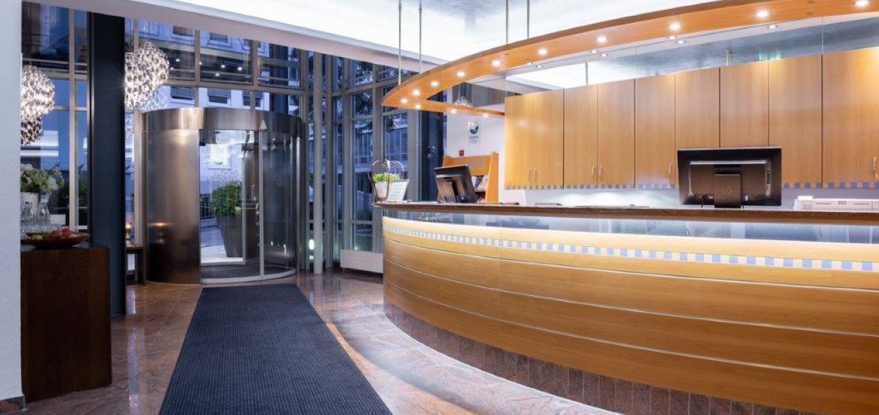 Modern hotel reception with curved wooden counter, illuminated reception area and revolving door in the background. Elegant, inviting ambience., © Kronen Hotel GmbH