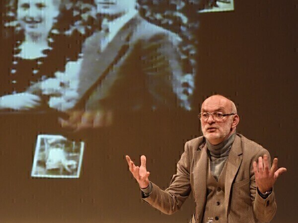 Ein Mann sitzt auf einer B&uuml;hne und gestikuliert. Im Hintergrund ist ein altes Schwarz-Wei&szlig;-Foto eines Paares projiziert., &copy; Theaterhaus Stuttgart e.V.