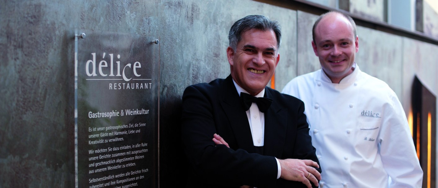 Zwei Männer stehen vor einem Schild des Délice Restaurants. Einer trägt einen Anzug, der andere eine Kochjacke., © Delice