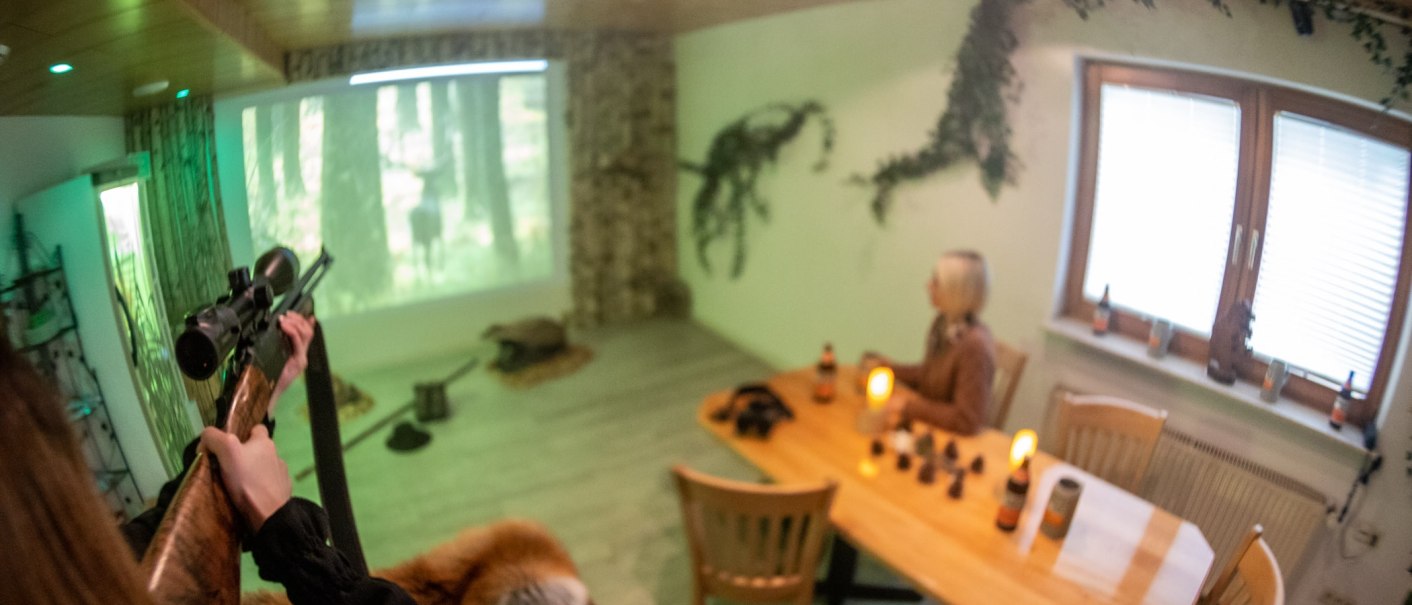 A room with hunting motifs. A person is aiming a rifle at a projection. Another person is sitting at a table with drinks and candles., &copy; TOMAS
