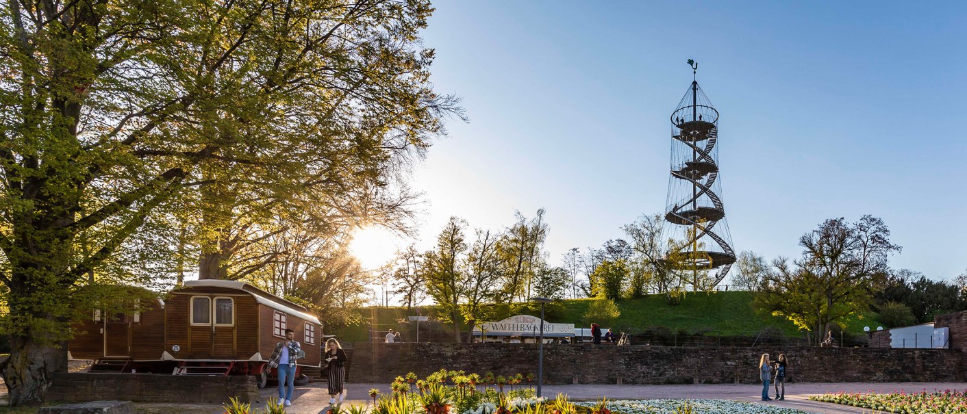 Der Killesbergturm steht in einem Park, umgeben von Bäumen. Menschen spazieren bei Sonnenuntergang. Ein Waffelstand ist sichtbar., © Stuttgart-Marketing GmbH, Werner Dieterich