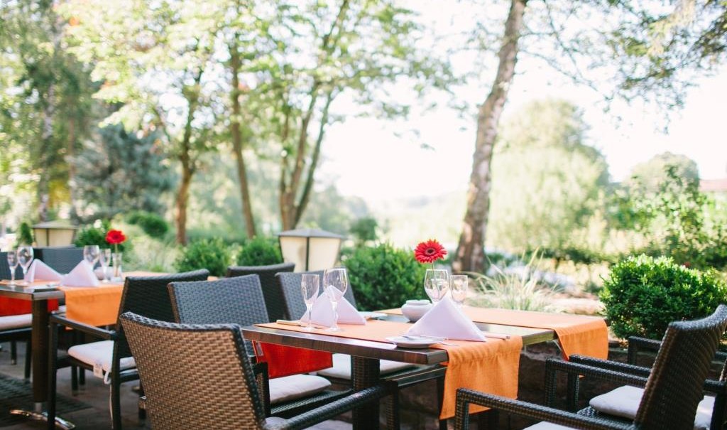 Tables laid out on a terrace with orange table runners and red flowers, surrounded by trees and shrubs., &copy; relexa Waldhotel Schatten