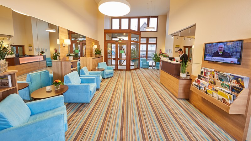 Helle Lobby mit blauen Sesseln, Holzmöbeln und gestreiftem Teppich. Rezeption mit Mitarbeiter, TV an der Wand, Zeitschriftenständer., © TOMAS Helle Lobby mit blauen Sesseln, Holzmöbeln und gestreiftem Teppich. Rezeption mit Mitarbeiter, TV an der Wand, Zeitschriftenständer., © TOMAS