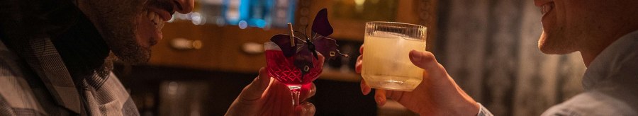 Two men clinking cocktails in a bar, one in a checked jacket, the other in a light-colored shirt. Bottles can be seen in the background., &copy; &copy; Stuttgart-Marketing GmbH, Martina Denker
