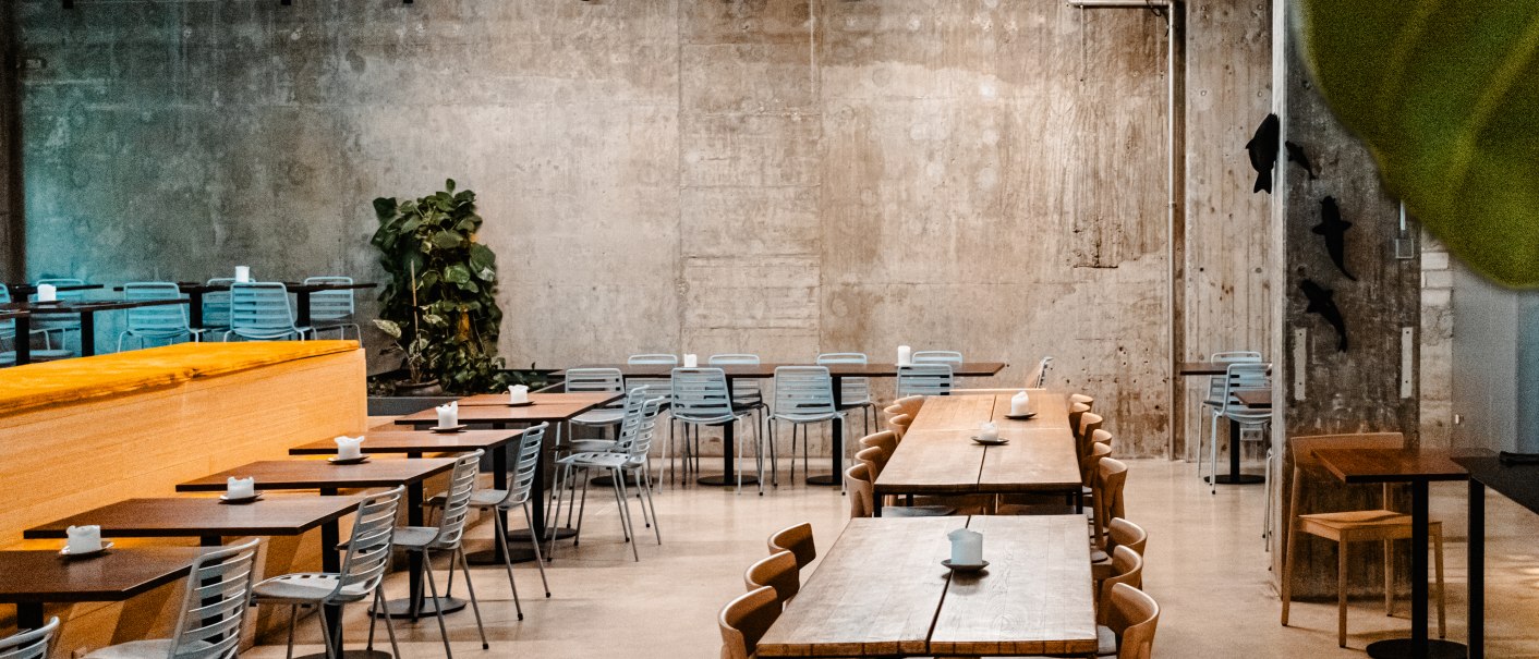 Leerer, moderner Cafébereich mit Holztischen und Metallstühlen vor einer Betonwand. Pflanzen und Beleuchtung sorgen für eine gemütliche Atmosphäre., © SMG Stuttgart Marketing GmbH - Sarah Schmid Leerer, moderner Cafébereich mit Holztischen und Metallstühlen vor einer Betonwand. Pflanzen und Beleuchtung sorgen für eine gemütliche Atmosphäre., © SMG Stuttgart Marketing GmbH - Sarah Schmid
