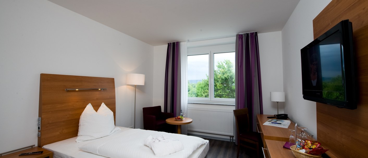 A cozy single room with a bed, desk, TV and window. There is an armchair and a small table next to the window., © Hotel Fortuna