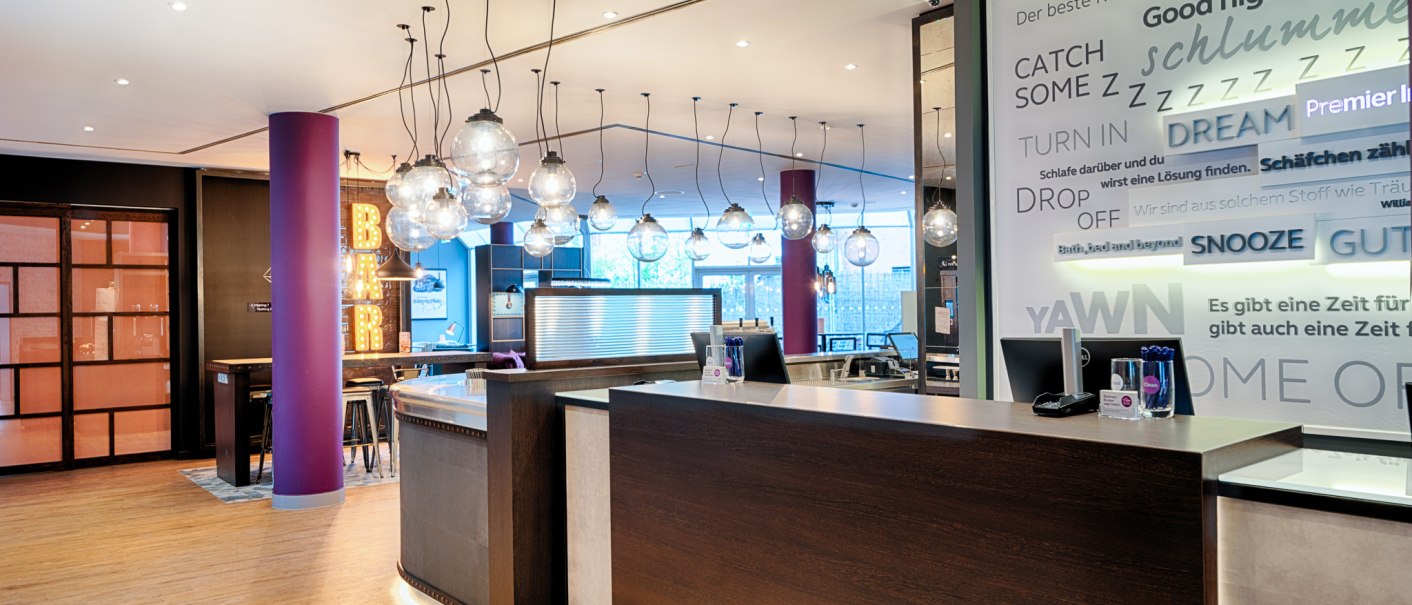 Moderne Hotelrezeption mit h&auml;ngenden Glaskugelleuchten. Im Hintergrund ist eine Bar mit leuchtendem Schriftzug zu sehen. Die W&auml;nde sind mit Texten dekoriert., &copy; Premier Inn Germany