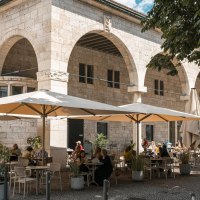 Ein Café mit großen Sonnenschirmen und Gästen, die draußen an Tischen sitzen. Das Gebäude hat Bögen und ist aus Stein., © Stuttgart Marketing GmbH, Sarah Schmid