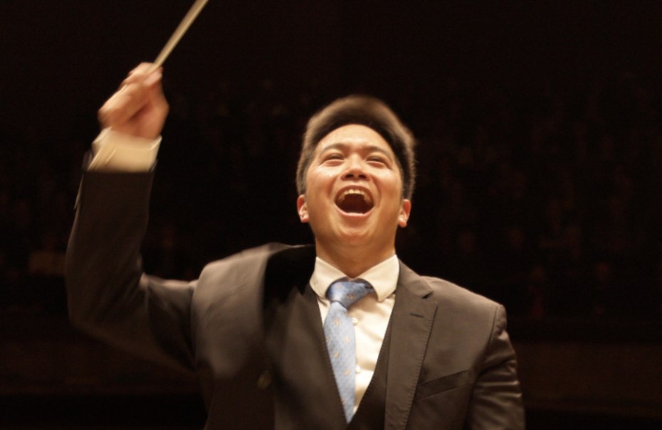 Ein l&auml;chelnder Dirigent in einem Anzug dirigiert mit einem Taktstock vor einem dunklen Hintergrund., &copy; Jochen Stall