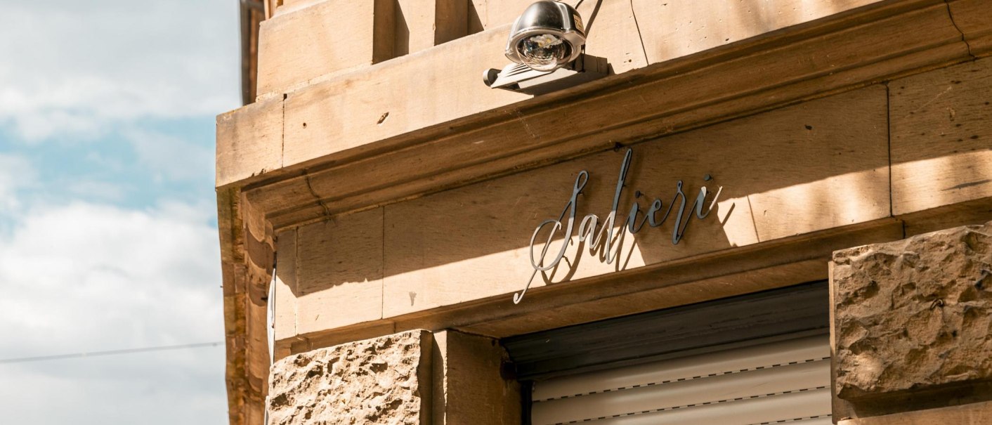 Fassade eines Geb&auml;udes mit dem Schriftzug 'Salieri' und einer &Uuml;berwachungskamera. Die Sonne wirft Schatten auf die sandfarbene Wand., &copy; SMG, Sarah Schmid