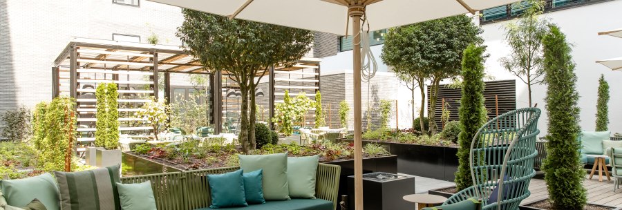 Moderne Terrasse mit gr&uuml;nen Sitzm&ouml;beln, gro&szlig;em Sonnenschirm und &uuml;ppiger Bepflanzung. Einladende Atmosph&auml;re mit Holzboden und stilvollen Details., &copy; Motel One - Stuttgart Hauptbahnhof