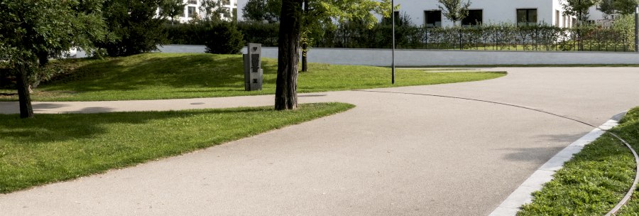 Ein gepflegter Park mit geschwungenen Gehwegen, Bäumen und modernen Wohngebäuden im Hintergrund. Ein Gedenkstein ist sichtbar., © Foto-Kunst Andreas Keller Ein gepflegter Park mit geschwungenen Gehwegen, Bäumen und modernen Wohngebäuden im Hintergrund. Ein Gedenkstein ist sichtbar., © Foto-Kunst Andreas Keller