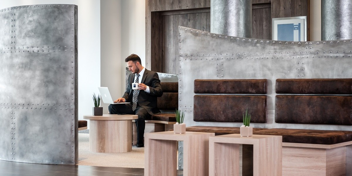A man in a suit is working in a modern lobby. He is holding a coffee cup and using a laptop. The furnishings are stylish and minimalist., &copy; Essential by Dorint