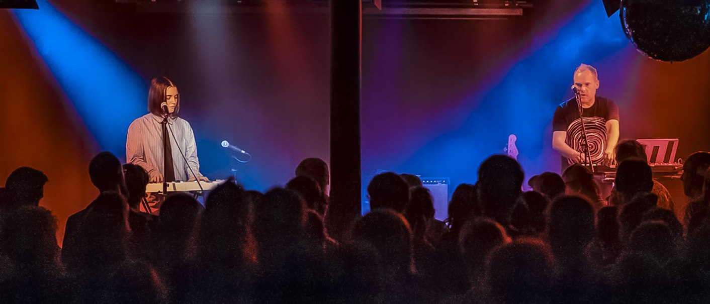 Two musicians on a stage play keyboards. Colorful lights in blue and red illuminate the scene. An audience looks on., © Martin Schniz Two musicians on a stage play keyboards. Colorful lights in blue and red illuminate the scene. An audience looks on., © Martin Schniz