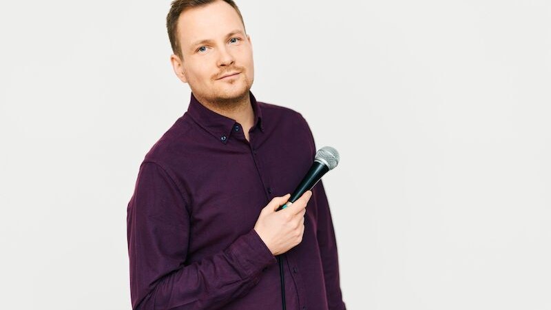 Ein Mann in einem lila Hemd hält ein Mikrofon und blickt leicht lächelnd in die Kamera. Der Hintergrund ist neutral und hell., © Rosenau Kultur e.V. Ein Mann in einem lila Hemd hält ein Mikrofon und blickt leicht lächelnd in die Kamera. Der Hintergrund ist neutral und hell., © Rosenau Kultur e.V.